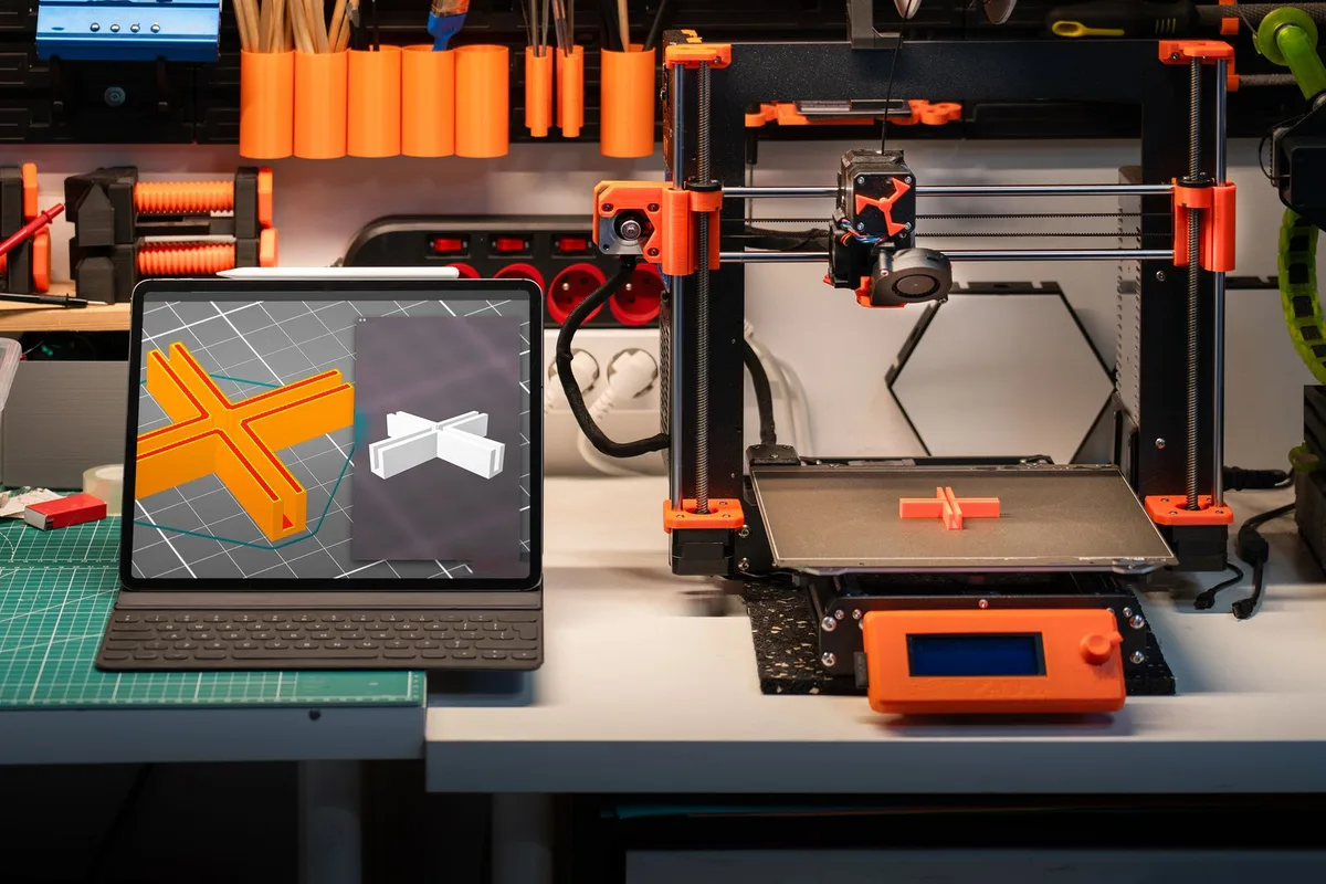 A beginner 3D printer workspace with an open-frame FDM printer mid-print next to a tablet showing the 3D model being printed