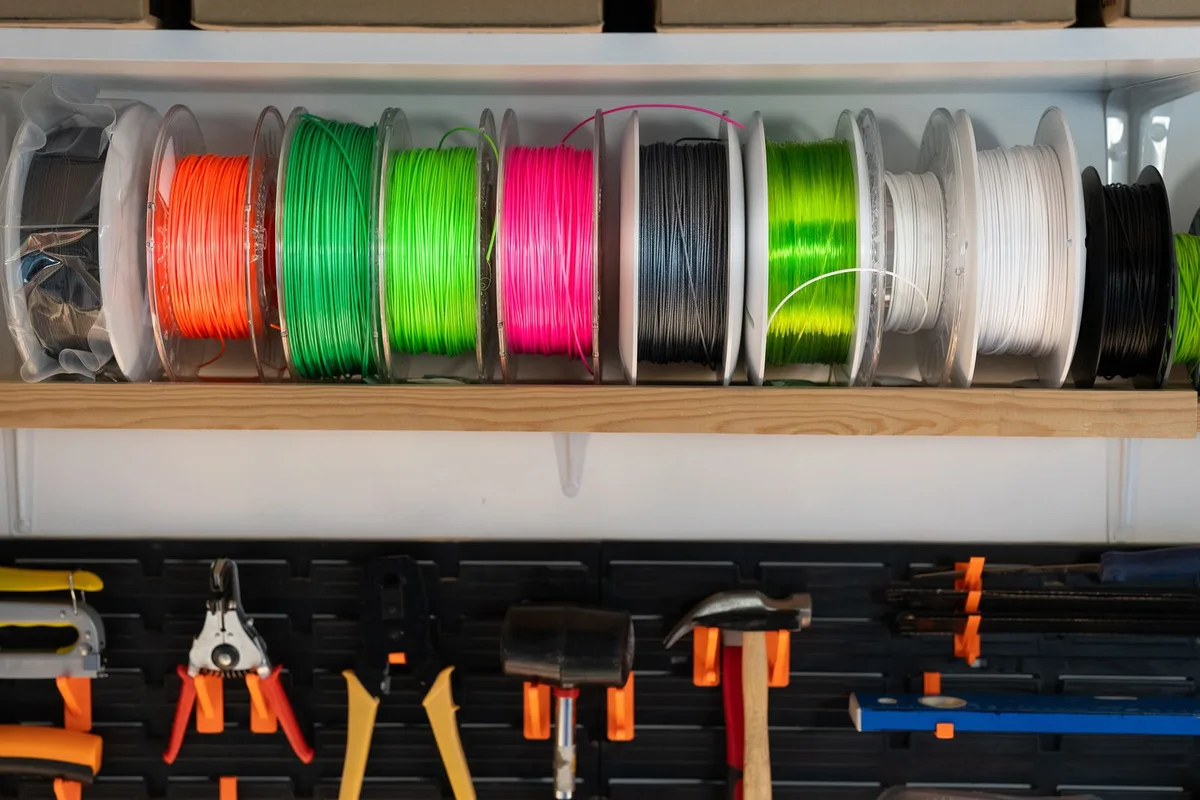 Shelf of colorful PLA, PETG, and ABS 3D printer filament spools in orange, green, pink, black, and white