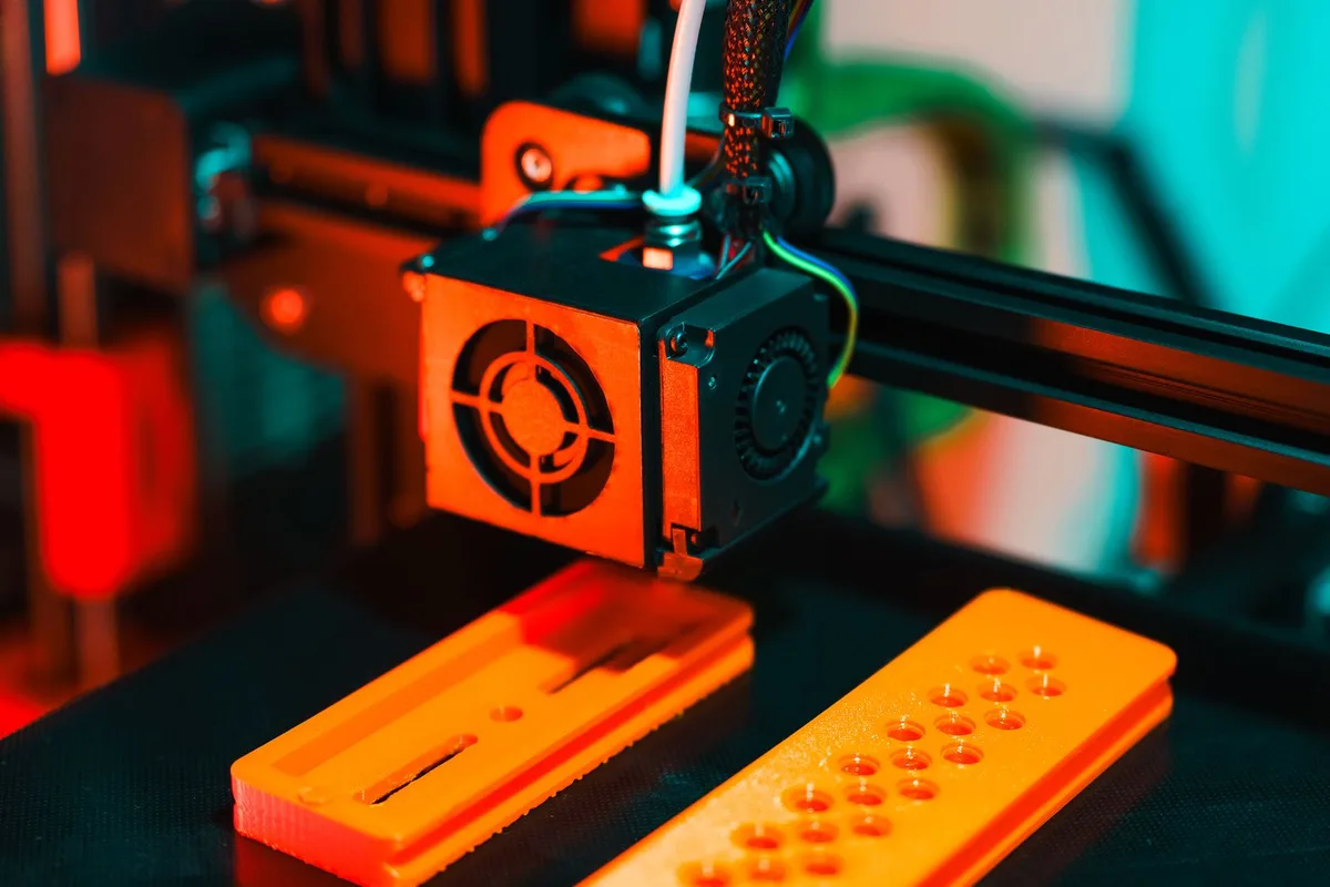 Close-up of a 3D printer hotend extruding orange plastic onto two finished parts